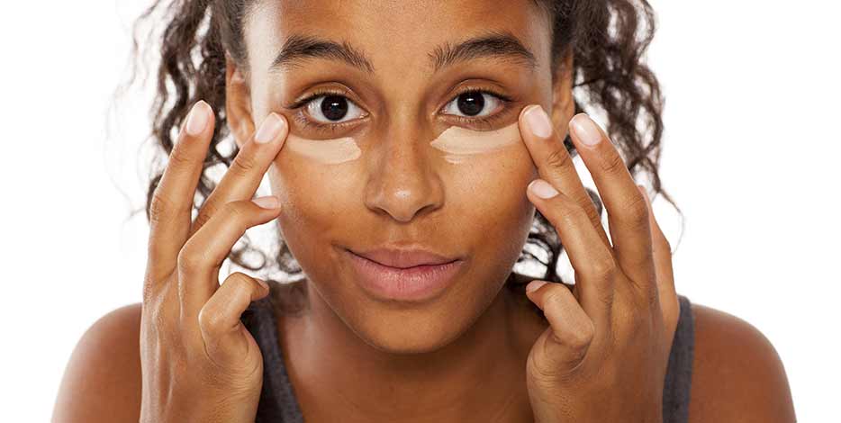 A younger woman applies concealer under her eyes with her fingers to hide dark circles under her eyes