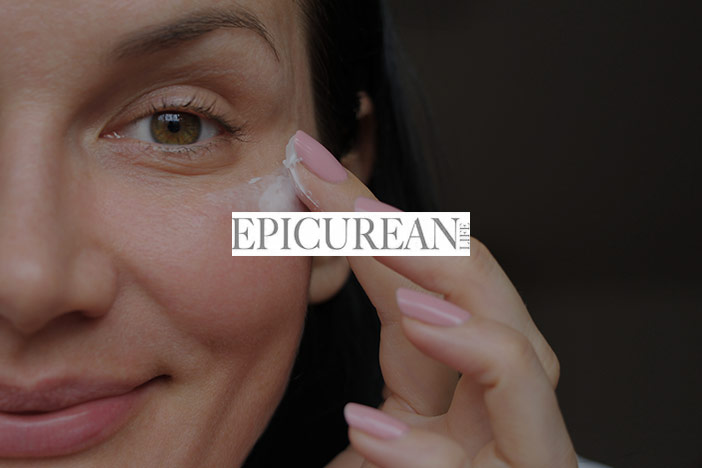 A middle aged woman applies cream to the eye area of her face. The Epicurean Life logo is superimposed on top.