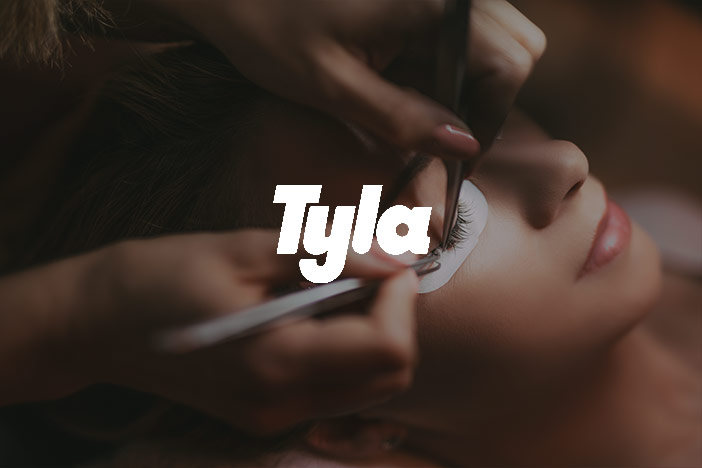 A middle aged woman is having eyelash extensions placed on by a lash technician with the Tyla logo superimposed on top