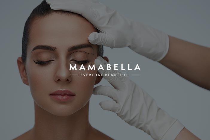 Doctor examining a woman's eyelid for blepharoplasty with Mamabella logo superimposed