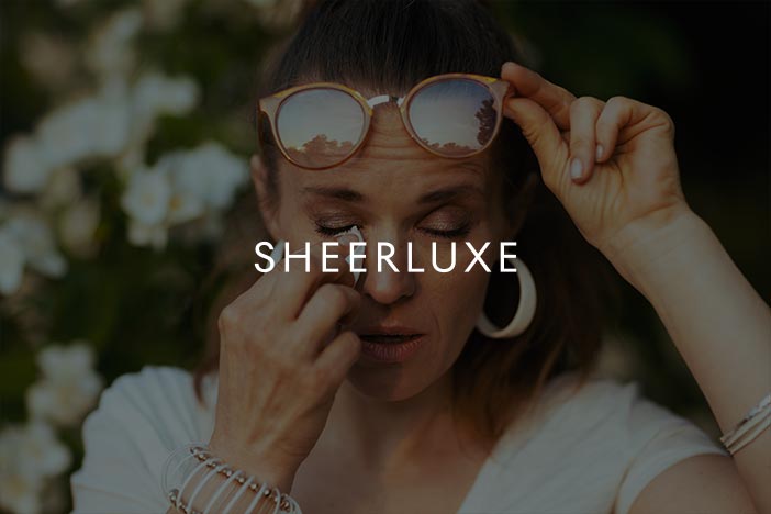 Woman Experiencing Hay Fever SheerLuxe Feature Image Woman rubbing her irritated eyes outdoors while wearing sunglasses on her head, representing hay fever symptoms.