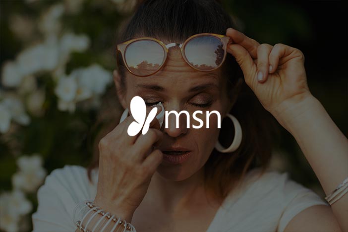 Woman outdoors wiping her eye with a tissue while lifting sunglasses, featured in MSN article about allergy symptoms