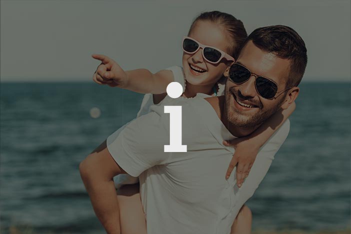 Father giving daughter a piggyback ride at the beach, both wearing sunglasses and smiling, with the ocean in the background.